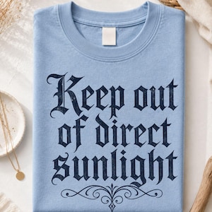 Op de afbeelding: Een lichtblauw t-shirt met ronde hals en de donker marineblauwe tekst "Keep out of direct sunlight" gedrukt in een gotisch lettertype, met een decoratieve krul onder de tekst en een klein "BLue"-logo bij de kraag.