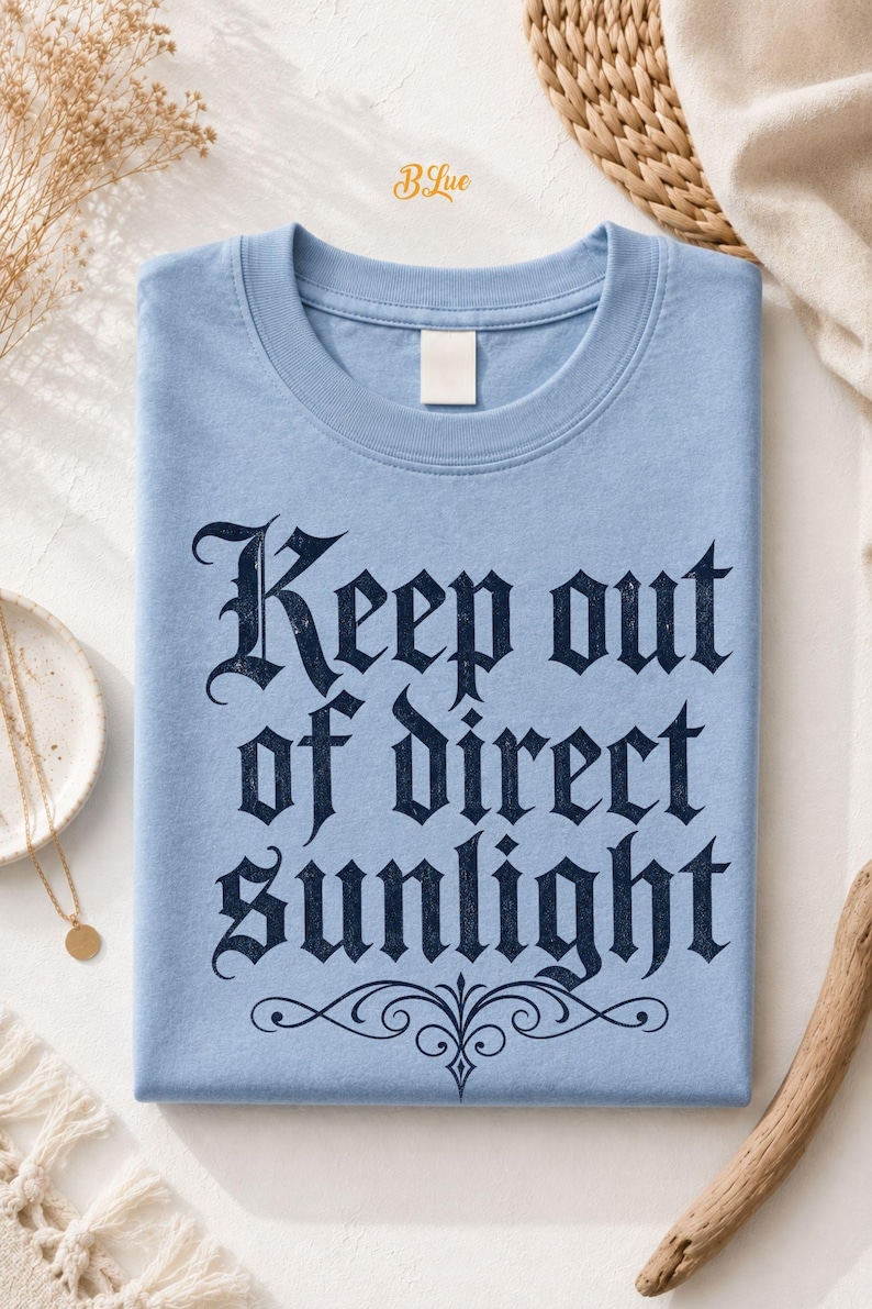 Op de afbeelding: Een lichtblauw t-shirt met ronde hals en de donker marineblauwe tekst "Keep out of direct sunlight" gedrukt in een gotisch lettertype, met een decoratieve krul onder de tekst en een klein "BLue"-logo bij de kraag.