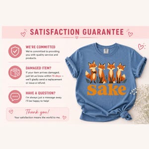 May include: A blue short-sleeved t-shirt featuring a graphic of four cartoon foxes standing above the orange text "sake". The shirt is displayed alongside text detailing a satisfaction guarantee, including policies on damaged items and customer support.