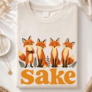 May include: A natural-colored t-shirt featuring a graphic of four orange and white foxes standing among green fern leaves. Below the foxes, the word "Sake" is printed in large, bold, orange letters. The shirt has a crew neckline and a plain label.