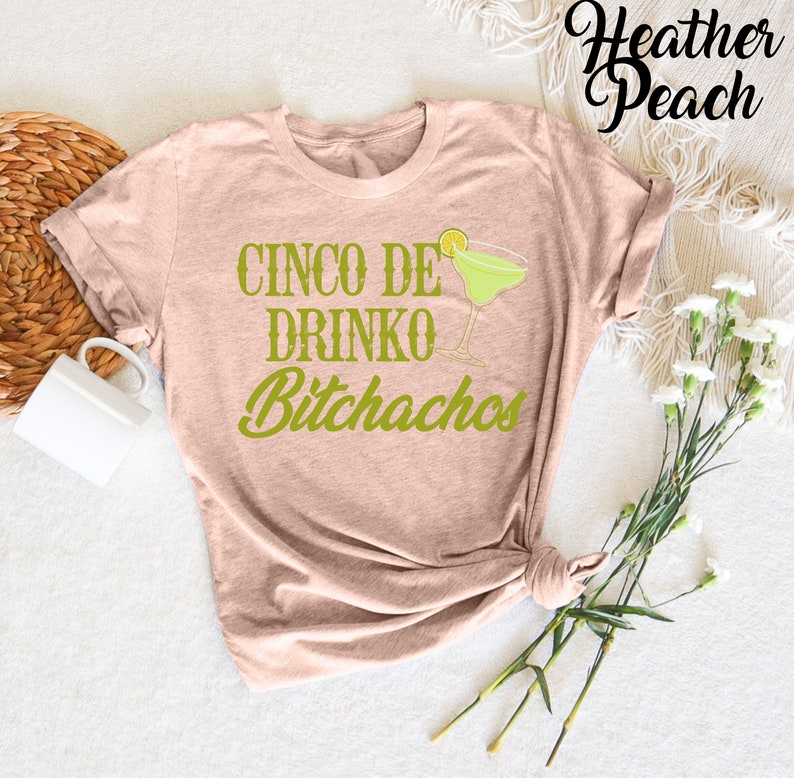 May include: A heather peach coloured t-shirt with the text "Cinco De Drinko Bitchachos" in green lettering. A green margarita glass with a lime wedge is pictured above the text.