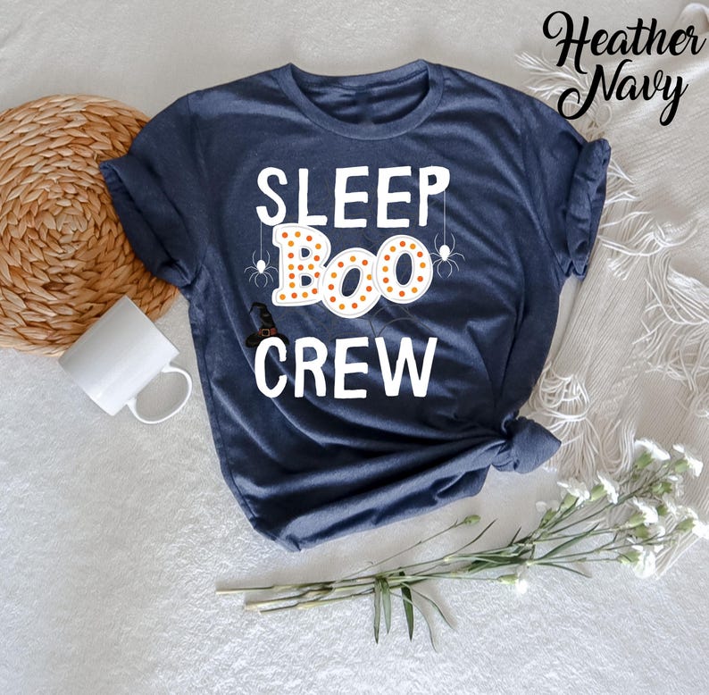 May include: A heather navy blue t-shirt with the text "SLEEP BOO CREW" in white. The word "BOO" is decorated with orange and yellow polka dots and surrounded by black spider webs.