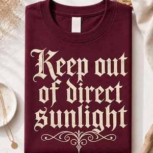 Op de afbeelding: Een bordeauxrood t-shirt met ronde hals en de cr&egrave;mekleurige tekst "Keep out of direct sunlight" in een gotisch lettertype, vergezeld van een decoratieve krul onder de tekst. Het shirt heeft een leeg wit label aan de binnenkant van de hals.