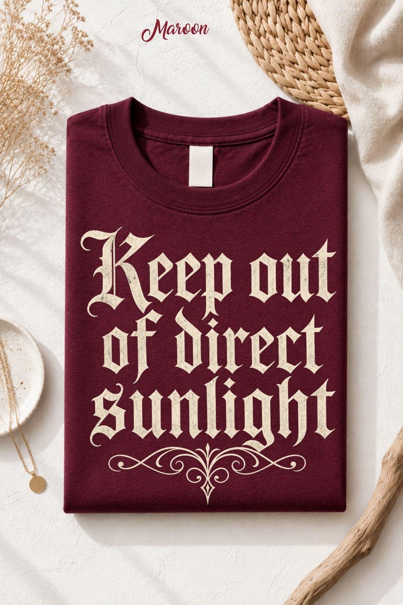 Op de afbeelding: Een bordeauxrood t-shirt met ronde hals en de cr&egrave;mekleurige tekst "Keep out of direct sunlight" in een gotisch lettertype, vergezeld van een decoratieve krul onder de tekst. Het shirt heeft een leeg wit label aan de binnenkant van de hals.