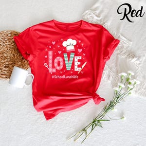 Pode incluir: Camiseta vermelha com um gr&aacute;fico da palavra "LOVE" com cora&ccedil;&otilde;es e um chap&eacute;u de cozinheiro. O texto "#SchoolLunchlife" est&aacute; abaixo do gr&aacute;fico.