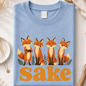 May include: A light blue t-shirt featuring a graphic of four orange foxes standing among green leaves. Below the foxes, the word "Sake" is printed in large, bold, orange letters. The shirt has a crew neckline and a plain, solid-colored back.