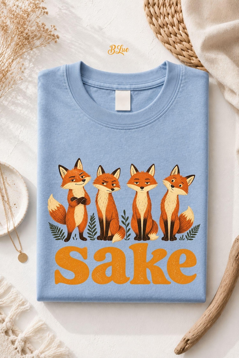 May include: A light blue t-shirt featuring a graphic of four orange foxes standing among green leaves. Below the foxes, the word "Sake" is printed in large, bold, orange letters. The shirt has a crew neckline and a plain, solid-colored back.