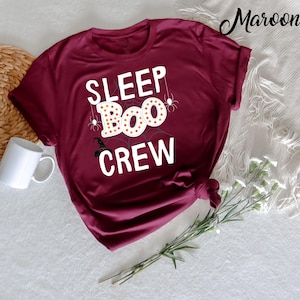May include: A maroon t-shirt with the text "SLEEP BOO CREW" in white letters. The text is surrounded by a spider web design with a witch's hat and spiders.