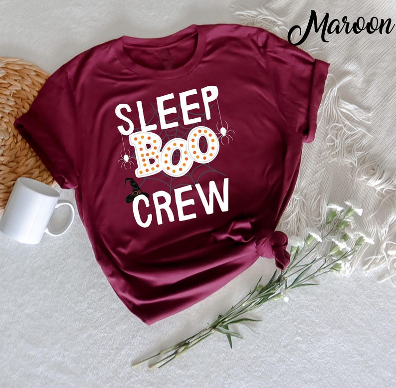 May include: A maroon t-shirt with the text "SLEEP BOO CREW" in white letters. The text is surrounded by a spider web design with a witch's hat and spiders.