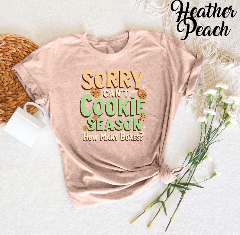 May include: A light brown t-shirt with the text "Sorry can't cookie season. How many boxes?" in green and brown. The text is surrounded by images of chocolate chip cookies.