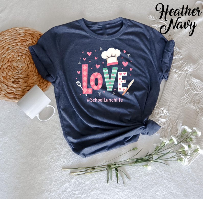 Pode incluir: Camiseta azul marinho heather com um gr&aacute;fico do texto "LOVE" em rosa e verde com um chap&eacute;u de cozinheiro, uma faca e uma esp&aacute;tula. O texto "#SchoolLunchlife" est&aacute; abaixo do gr&aacute;fico.