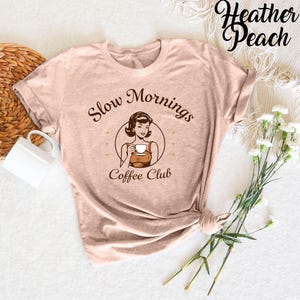 Puede incluir: Una camiseta de color melocot&oacute;n brezo con una ilustraci&oacute;n retro de una mujer tomando caf&eacute;. El texto de la camiseta dice "Slow Mornings Coffee Club".