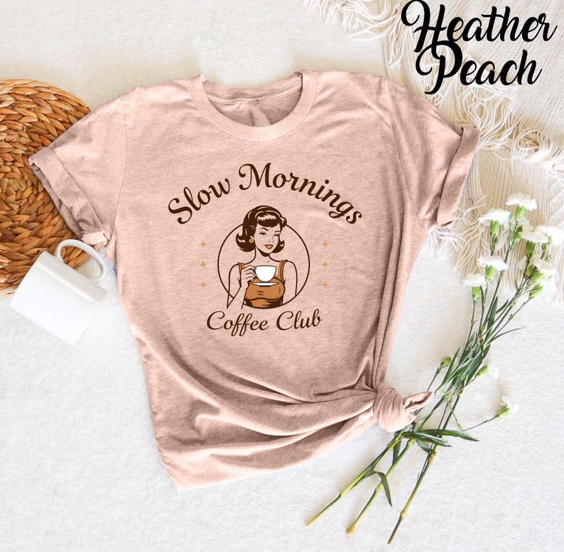 Puede incluir: Una camiseta de color melocot&oacute;n brezo con una ilustraci&oacute;n retro de una mujer tomando caf&eacute;. El texto de la camiseta dice "Slow Mornings Coffee Club".