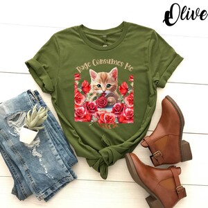 May include: Olive green t-shirt featuring a graphic of a kitten surrounded by red roses, with the text "Rage Consumes Me." The shirt is paired with distressed blue jeans and brown ankle boots.