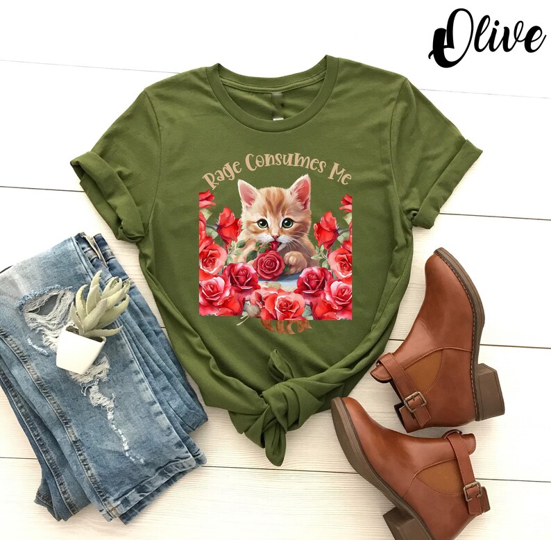May include: Olive green t-shirt featuring a graphic of a kitten surrounded by red roses, with the text "Rage Consumes Me." The shirt is paired with distressed blue jeans and brown ankle boots.