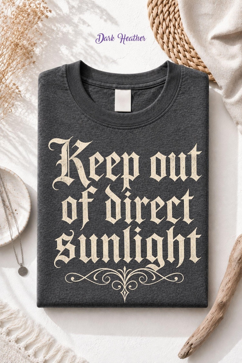 Op de afbeelding: Een donkergrijs gem&ecirc;leerd T-shirt met ronde hals en de tekst "Keep out of direct sunlight" gedrukt in een cr&egrave;mekleurig gotisch lettertype. Onder de tekst staat een decoratief krulontwerp. Het T-shirt heeft een leeg wit label in de nek.