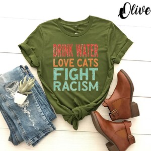 May include: Olive green t-shirt with the text "Drink Water Love Cats Fight Racism" in a rainbow color scheme.