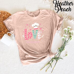 Pode incluir: Uma camiseta cor p&ecirc;ssego com um design gr&aacute;fico da palavra "LOVE" em rosa, verde e branco com cora&ccedil;&otilde;es e um chap&eacute;u de cozinheiro. O texto "Food Love Life" est&aacute; abaixo da palavra "LOVE".