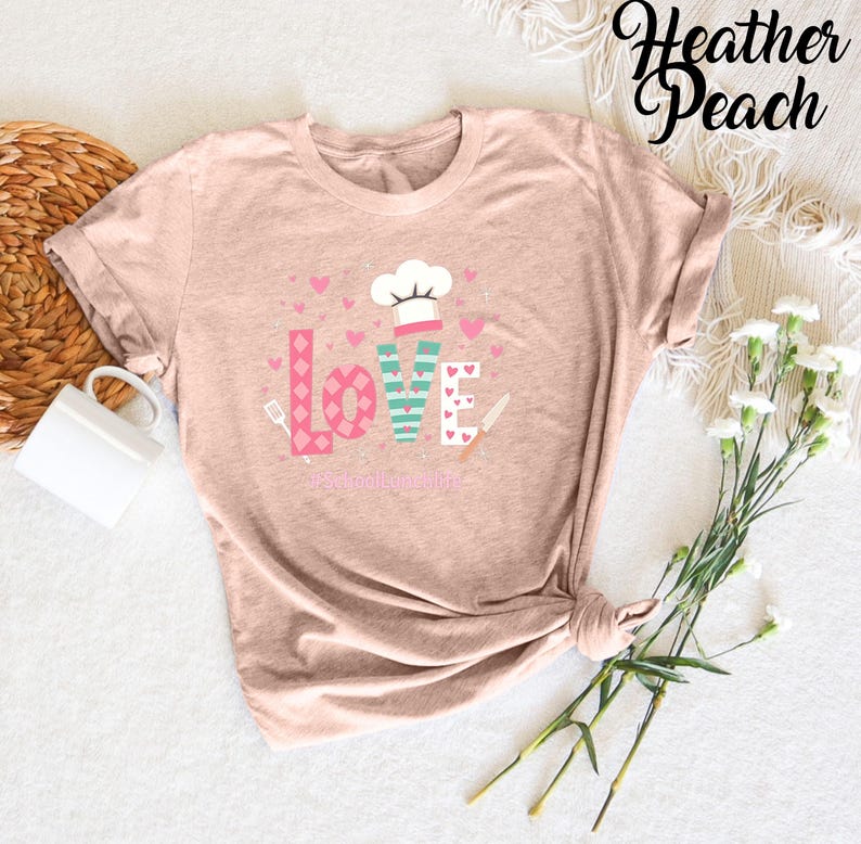 Pode incluir: Uma camiseta cor p&ecirc;ssego com um design gr&aacute;fico da palavra "LOVE" em rosa, verde e branco com cora&ccedil;&otilde;es e um chap&eacute;u de cozinheiro. O texto "Food Love Life" est&aacute; abaixo da palavra "LOVE".