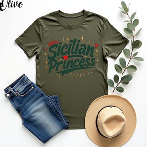 以下が含まれることがあります： 緑色のTシャツに「シチリアのプリンセス」という文字が緑色、赤色、金色のフォントで書かれています。文字の周りは小さな金色の星と赤いハートで囲まれています。