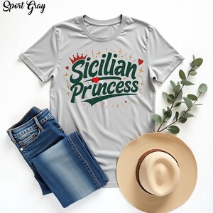 以下が含まれることがあります： 白い背景に置かれたグレーのTシャツ。Tシャツには緑と赤の文字で「シシリアン プリンセス」と書いてあり、王冠とハートが描かれている。Tシャツの隣には青いジーンズと麦わら帽子が置かれている。