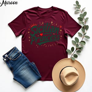 以下が含まれることがあります： 赤ワイン色のTシャツに、緑と金のグラフィックで「シチリアのプリンセス」と赤いハートが書かれています。