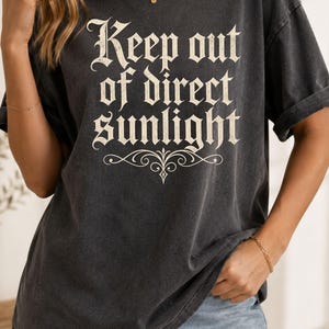 Op de afbeelding: Een antracietgrijs T-shirt met korte mouwen en de tekst Keep out of direct sunlight in een cr&egrave;mekleurig gotisch lettertype, vergezeld van een decoratief krulontwerp dat zich onder de tekst op de voorzijde van het kledingstuk bevindt.