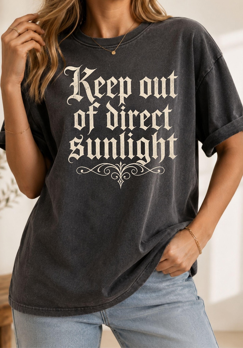 Op de afbeelding: Een antracietgrijs T-shirt met korte mouwen en de tekst Keep out of direct sunlight in een cr&egrave;mekleurig gotisch lettertype, vergezeld van een decoratief krulontwerp dat zich onder de tekst op de voorzijde van het kledingstuk bevindt.