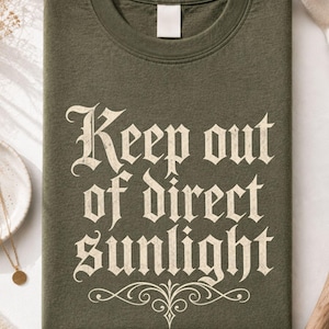 Op de afbeelding: Een olijfgroen t-shirt met ronde hals en de tekst "Keep out of direct sunlight" gedrukt in een verweerd cr&egrave;mekleurig gotisch lettertype, met een decoratief krul-element direct onder de tekst op de voorzijde van het kledingstuk.