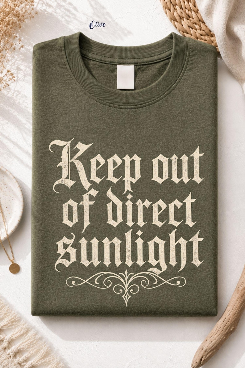 Op de afbeelding: Een olijfgroen t-shirt met ronde hals en de tekst "Keep out of direct sunlight" gedrukt in een verweerd cr&egrave;mekleurig gotisch lettertype, met een decoratief krul-element direct onder de tekst op de voorzijde van het kledingstuk.