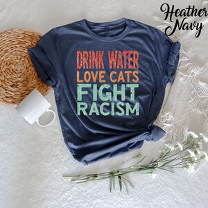 May include: A heather navy blue t-shirt with the text "Drink Water Love Cats Fight Racism" in a retro style font.
