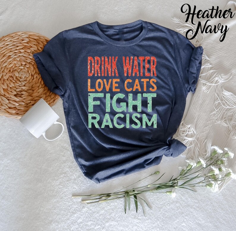 May include: A heather navy blue t-shirt with the text "Drink Water Love Cats Fight Racism" in a retro style font.
