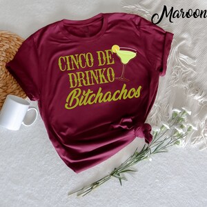 May include: A maroon t-shirt with the text "Cinco De Drinko Bitchachos" in gold lettering. A green margarita glass with a lime wedge is next to the text.