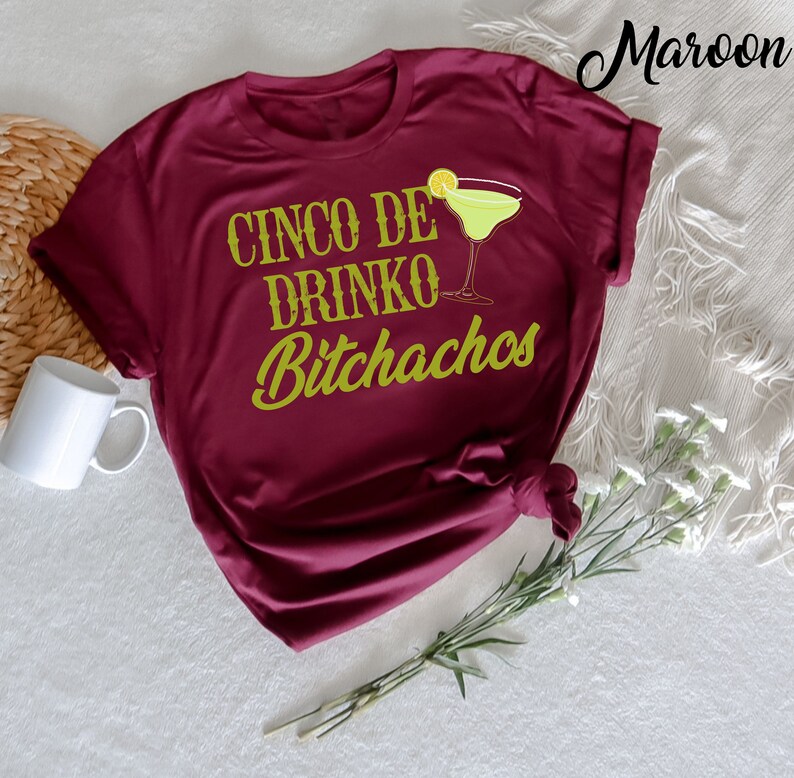May include: A maroon t-shirt with the text "Cinco De Drinko Bitchachos" in gold lettering. A green margarita glass with a lime wedge is next to the text.
