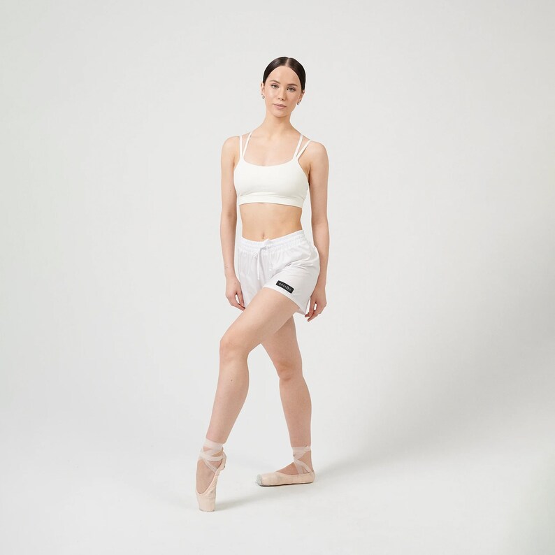 Puede incluir: Una mujer con un sujetador deportivo blanco y pantalones cortos blancos con un logotipo negro. Lleva zapatillas de ballet rosas y est&aacute; de pie con una pierna levantada.