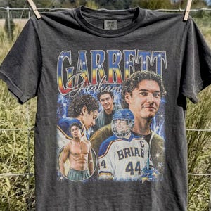 May include: Pepper-colored t-shirt featuring a vintage-style design with the name "Garrett Graham" in a multi-colored, retro font. The shirt includes multiple images of a person, with a hockey jersey and the number 44.