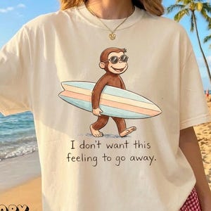 May include: Ivory-colored t-shirt featuring a cartoon monkey wearing sunglasses and carrying a surfboard. The shirt has the text "I don't want this feeling to go away." The word "IVORY" is printed at the bottom left.