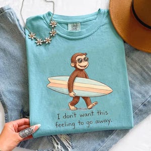 May include: A light blue Comfort Colors t-shirt featuring a cartoon monkey wearing sunglasses and carrying a surfboard. The shirt has the text "I don't want this feeling to go away." A necklace and a hat are also visible.