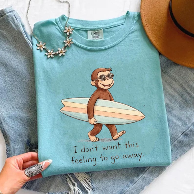 May include: A light blue Comfort Colors t-shirt featuring a cartoon monkey wearing sunglasses and carrying a surfboard. The shirt has the text "I don't want this feeling to go away." A necklace and a hat are also visible.