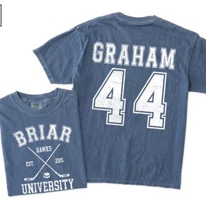 Puede incluir: Una camiseta color azul jean con gr&aacute;ficos serigrafiados en blanco. El frente muestra el texto BRIAR UNIVERSITY, HAWKS, EST. 2015 y dos palos de hockey cruzados con un disco. La parte posterior muestra el nombre GRAHAM y el n&uacute;mero 44.
