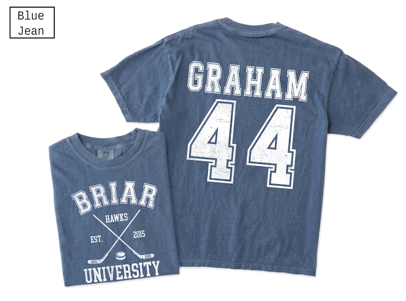 Puede incluir: Una camiseta color azul jean con gr&aacute;ficos serigrafiados en blanco. El frente muestra el texto BRIAR UNIVERSITY, HAWKS, EST. 2015 y dos palos de hockey cruzados con un disco. La parte posterior muestra el nombre GRAHAM y el n&uacute;mero 44.