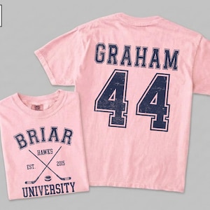Puede incluir: Una camiseta de manga corta de color rosa claro con gr&aacute;ficos azul oscuro de estilo universitario. El frente muestra 'BRIAR UNIVERSITY', 'HAWKS', 'EST. 2015' y palos de hockey cruzados con un disco. La parte posterior muestra el nombre 'GRAHAM' y el n&uacute;mero '44'.