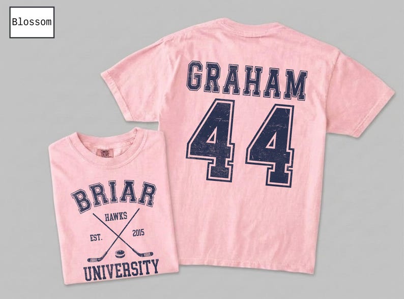 Puede incluir: Una camiseta de manga corta de color rosa claro con gr&aacute;ficos azul oscuro de estilo universitario. El frente muestra 'BRIAR UNIVERSITY', 'HAWKS', 'EST. 2015' y palos de hockey cruzados con un disco. La parte posterior muestra el nombre 'GRAHAM' y el n&uacute;mero '44'.