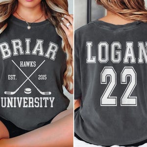 Może przedstawiać: Ciemnoszara koszulka z białym napisem. Z przodu napis "BRIAR HAWKS UNIVERSITY" ze skrzyżowanymi kijami hokejowymi i krążkiem. Z tyłu napis "LOGAN 22". Koszulka w stylu vintage, prawdopodobnie dla fana sportu.