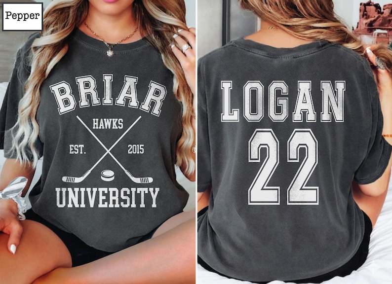 Może przedstawiać: Ciemnoszara koszulka z białym napisem. Z przodu napis "BRIAR HAWKS UNIVERSITY" ze skrzyżowanymi kijami hokejowymi i krążkiem. Z tyłu napis "LOGAN 22". Koszulka w stylu vintage, prawdopodobnie dla fana sportu.
