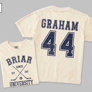 Puede incluir: Una camiseta de manga corta color marfil con un gr&aacute;fico azul marino en la parte delantera con el texto BRIAR UNIVERSITY, HAWKS, EST. 2015 y palos de hockey cruzados. La parte trasera muestra el nombre GRAHAM y el n&uacute;mero 44 en letra universitaria.