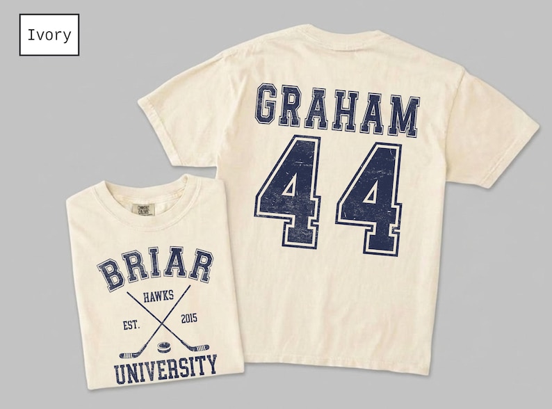Puede incluir: Una camiseta de manga corta color marfil con un gr&aacute;fico azul marino en la parte delantera con el texto BRIAR UNIVERSITY, HAWKS, EST. 2015 y palos de hockey cruzados. La parte trasera muestra el nombre GRAHAM y el n&uacute;mero 44 en letra universitaria.