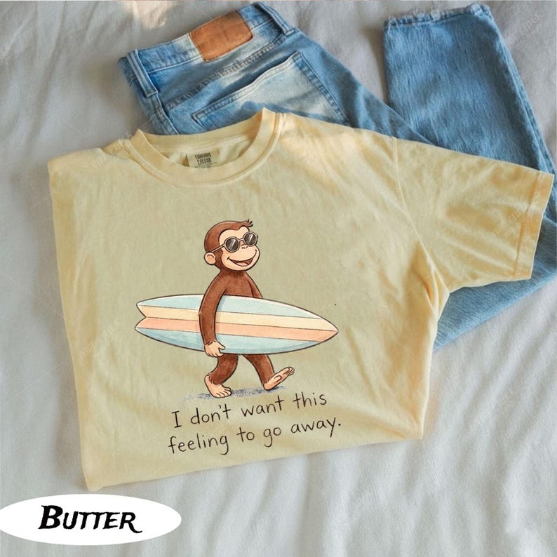 May include: Yellow t-shirt featuring a cartoon monkey wearing sunglasses and carrying a surfboard. The shirt has the text "I don't want this feeling to go away." Light blue jeans are folded next to the shirt. The word "Butter" is at the bottom.