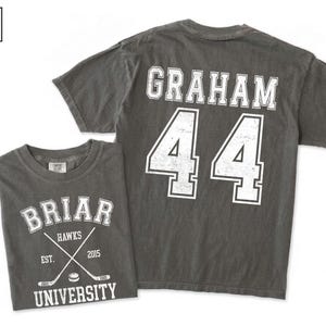 Puede incluir: Una camiseta de manga corta color pimienta con gr&aacute;ficos serigrafiados en blanco. El frente muestra 'BRIAR UNIVERSITY', 'HAWKS', 'EST. 2015' y palos de hockey cruzados. La parte posterior presenta el nombre 'GRAHAM' sobre el n&uacute;mero '44'.