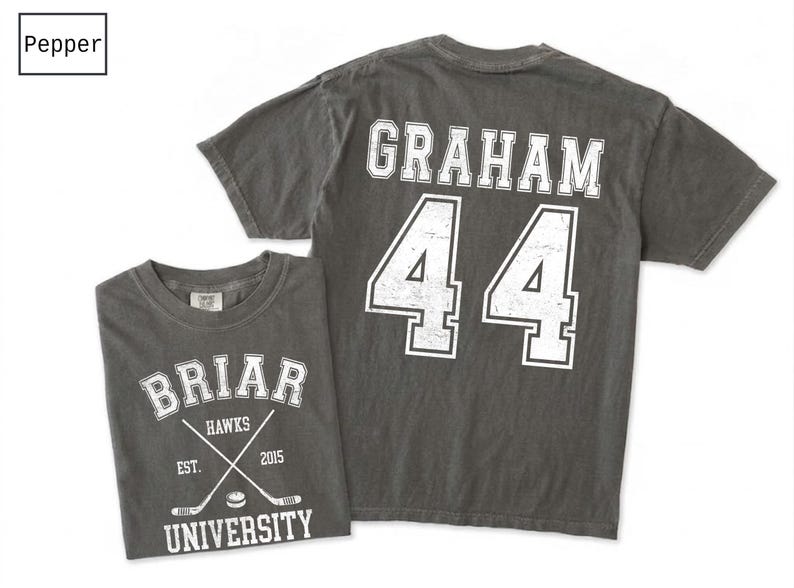 Puede incluir: Una camiseta de manga corta color pimienta con gr&aacute;ficos serigrafiados en blanco. El frente muestra 'BRIAR UNIVERSITY', 'HAWKS', 'EST. 2015' y palos de hockey cruzados. La parte posterior presenta el nombre 'GRAHAM' sobre el n&uacute;mero '44'.
